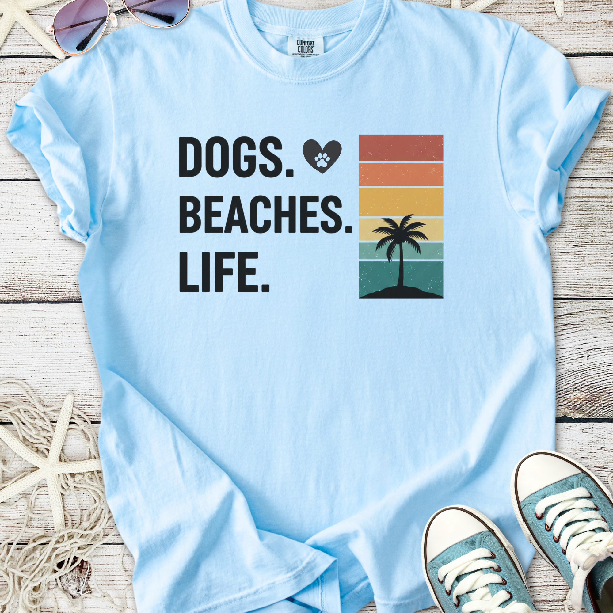 Light blue t-shirt with "Dogs. Beaches. Life." graphic, featuring a palm tree and sunset colors.