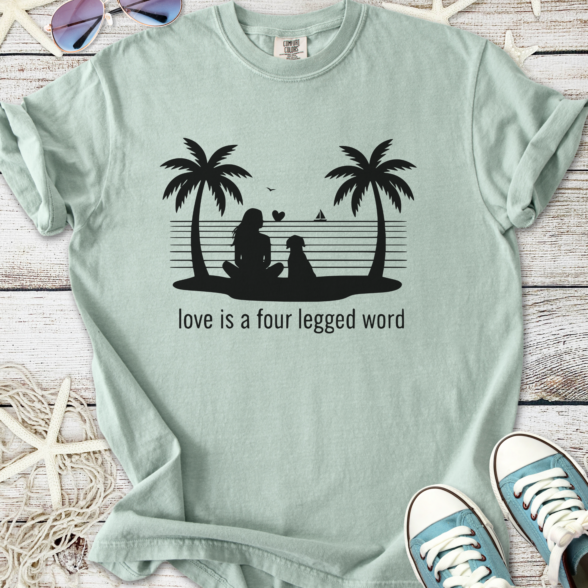 Mint green t-shirt with printed design of a woman and dog under palm trees with slogan about love.