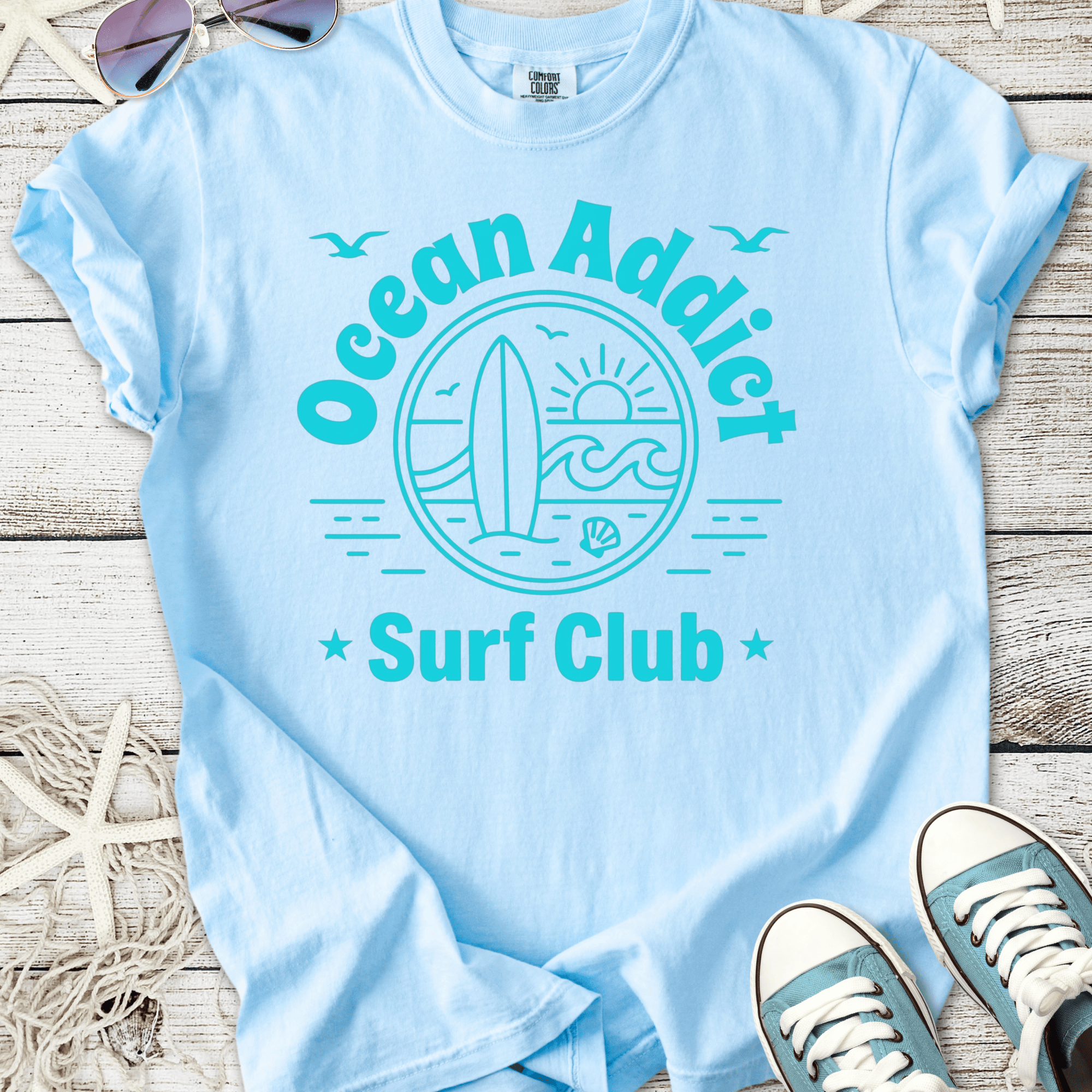 Light blue Ocean Addict Surf Club t-shirt featuring surfboard and sun graphic, perfect for beach lovers.