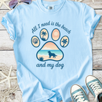 Light blue t-shirt featuring beach-themed graphic with paw print, text, and dog silhouette, perfect for dog lovers.