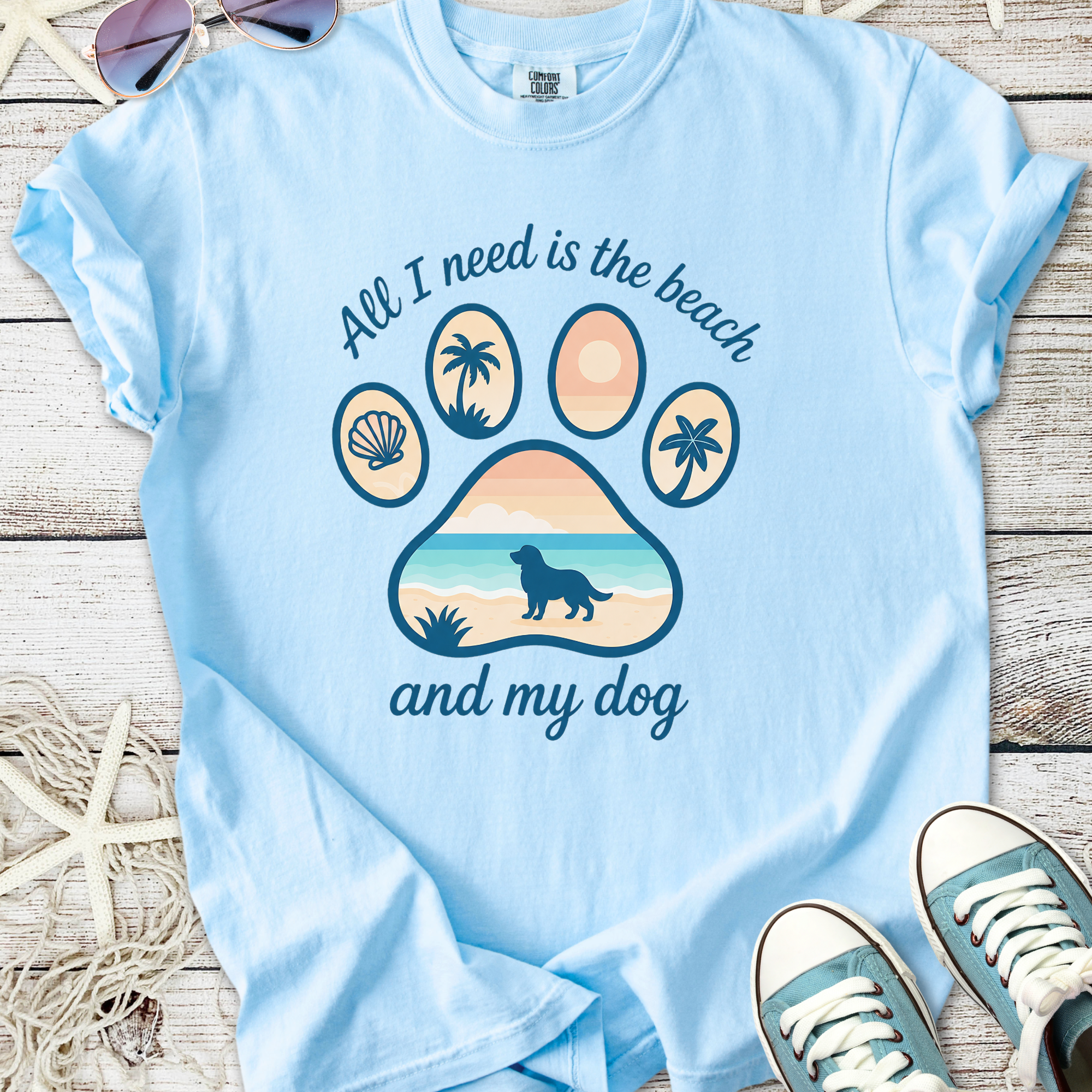 Light blue t-shirt featuring beach-themed graphic with paw print, text, and dog silhouette, perfect for dog lovers.