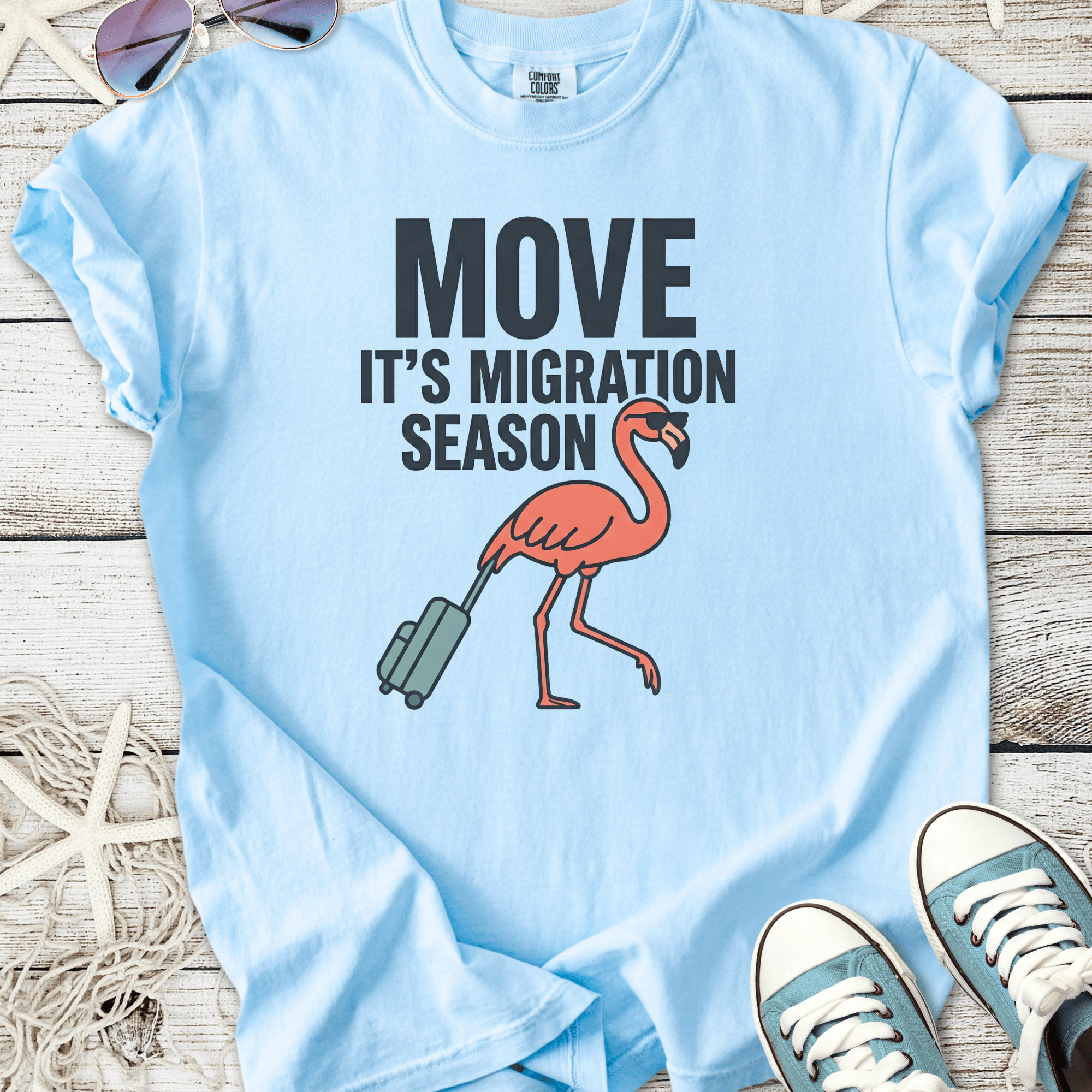 Light blue t-shirt featuring a flamingo with luggage and 'Move It's Migration Season' text.