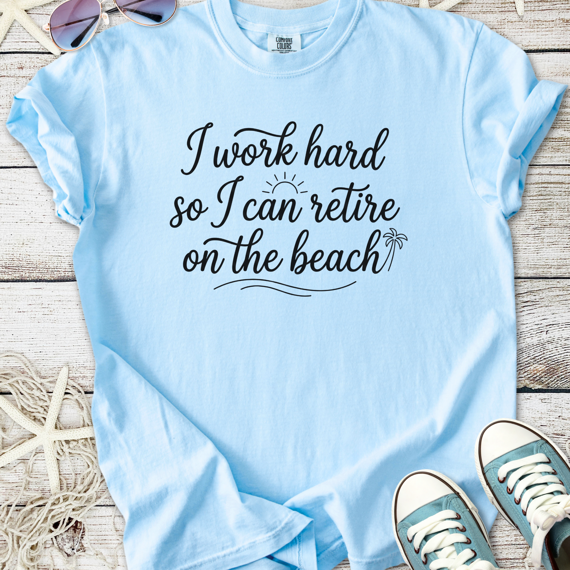 Light blue graphic tee with 'I work hard so I can retire on the beach' text, perfect for beach lovers.