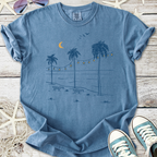 Light blue graphic t-shirt featuring palm trees, beach scene, and string lights for a casual summer vibe.
