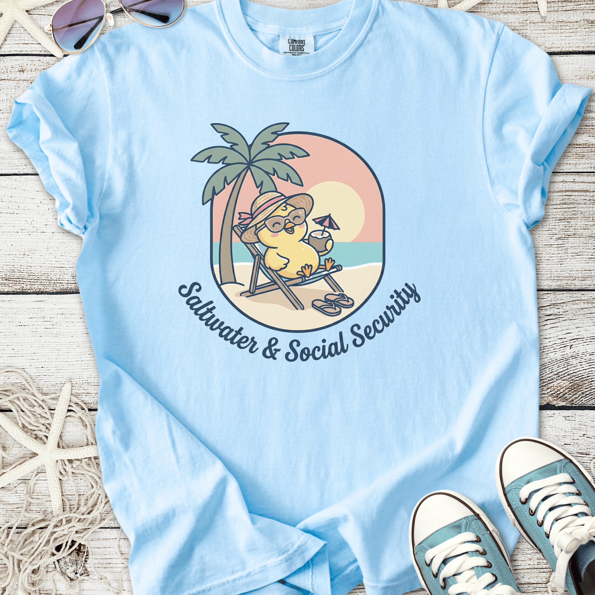 Light blue graphic tee featuring a relaxed cartoon character with sunglasses and cocktail, slogan 'Saltwater & Social Security'.