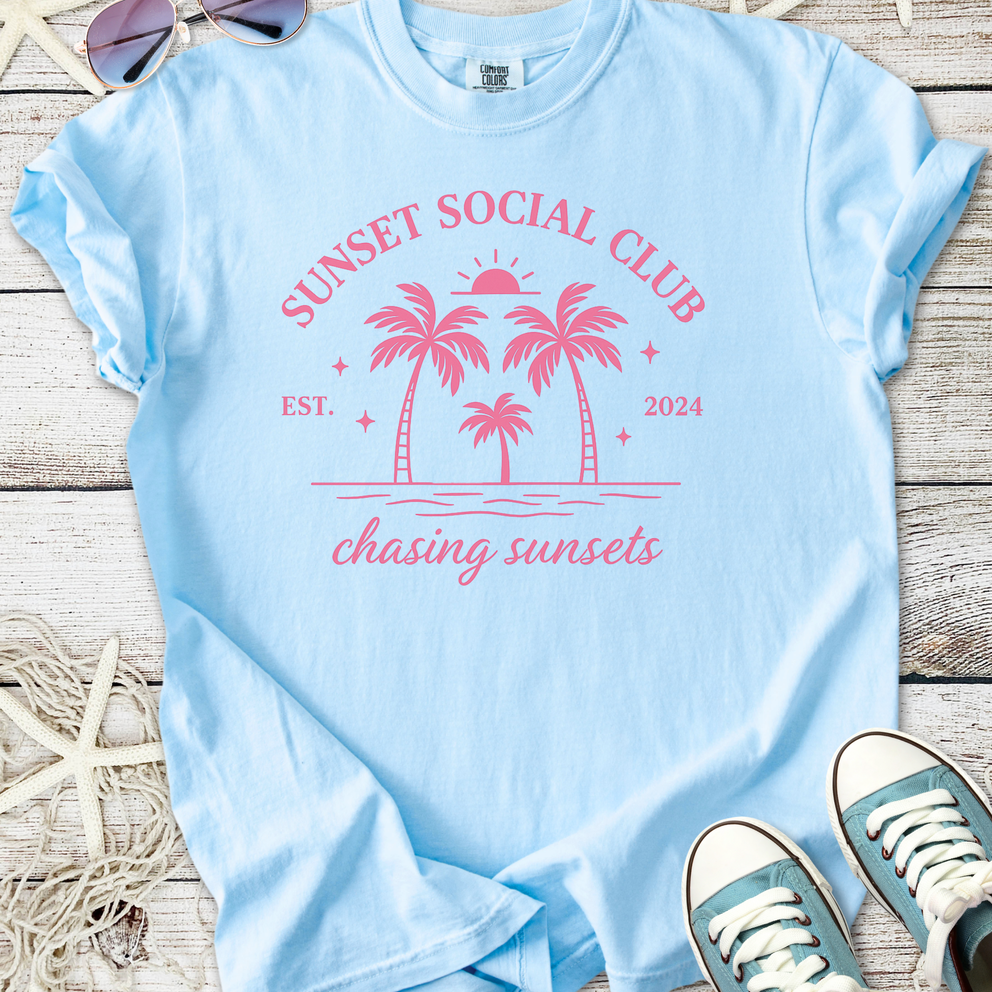 Light blue t-shirt featuring 'Sunset Social Club' print with palm trees, perfect for beach lovers.