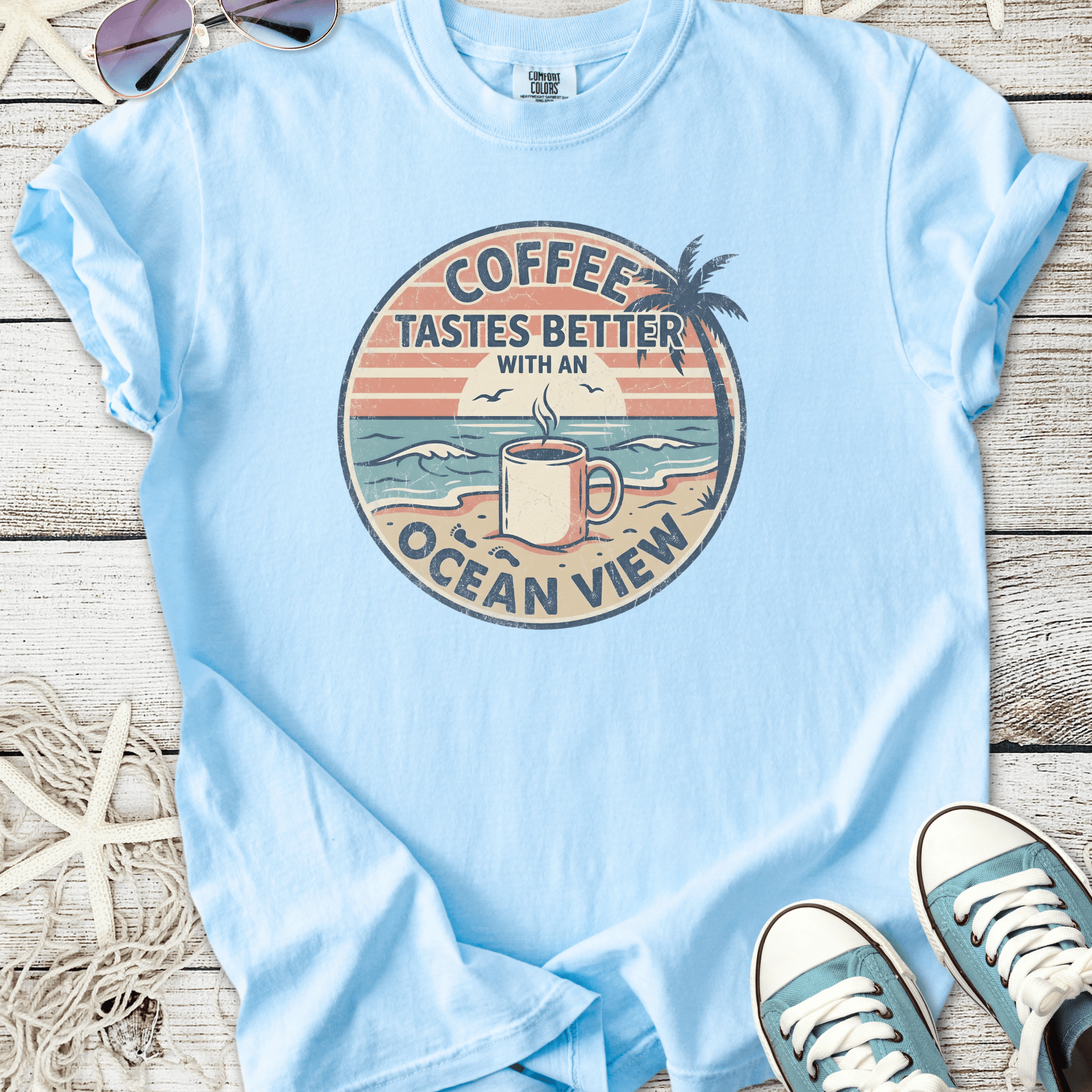 Light blue t-shirt featuring 'Coffee Tastes Better with an Ocean View' graphic, perfect for beach lovers.