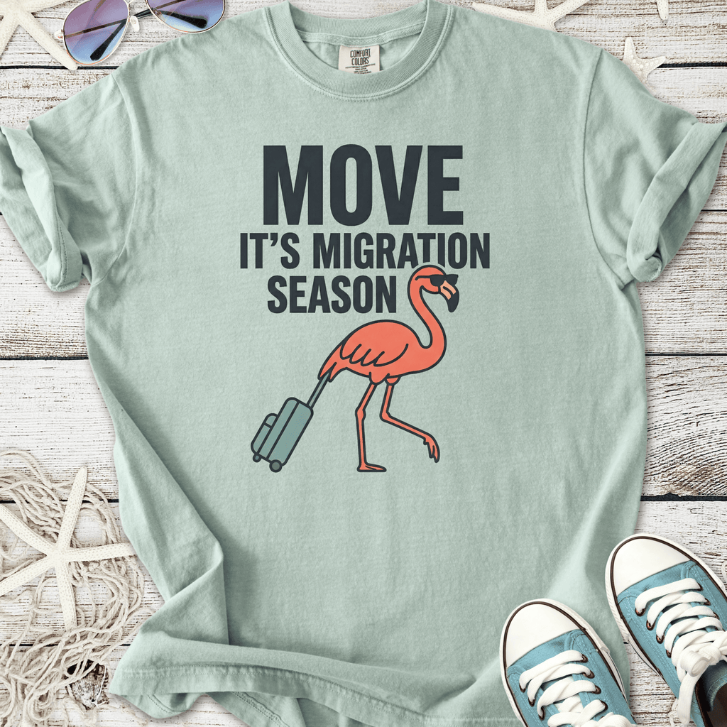 Light teal t-shirt with 'Move It's Migration Season' graphic and a pink flamingo carrying luggage.