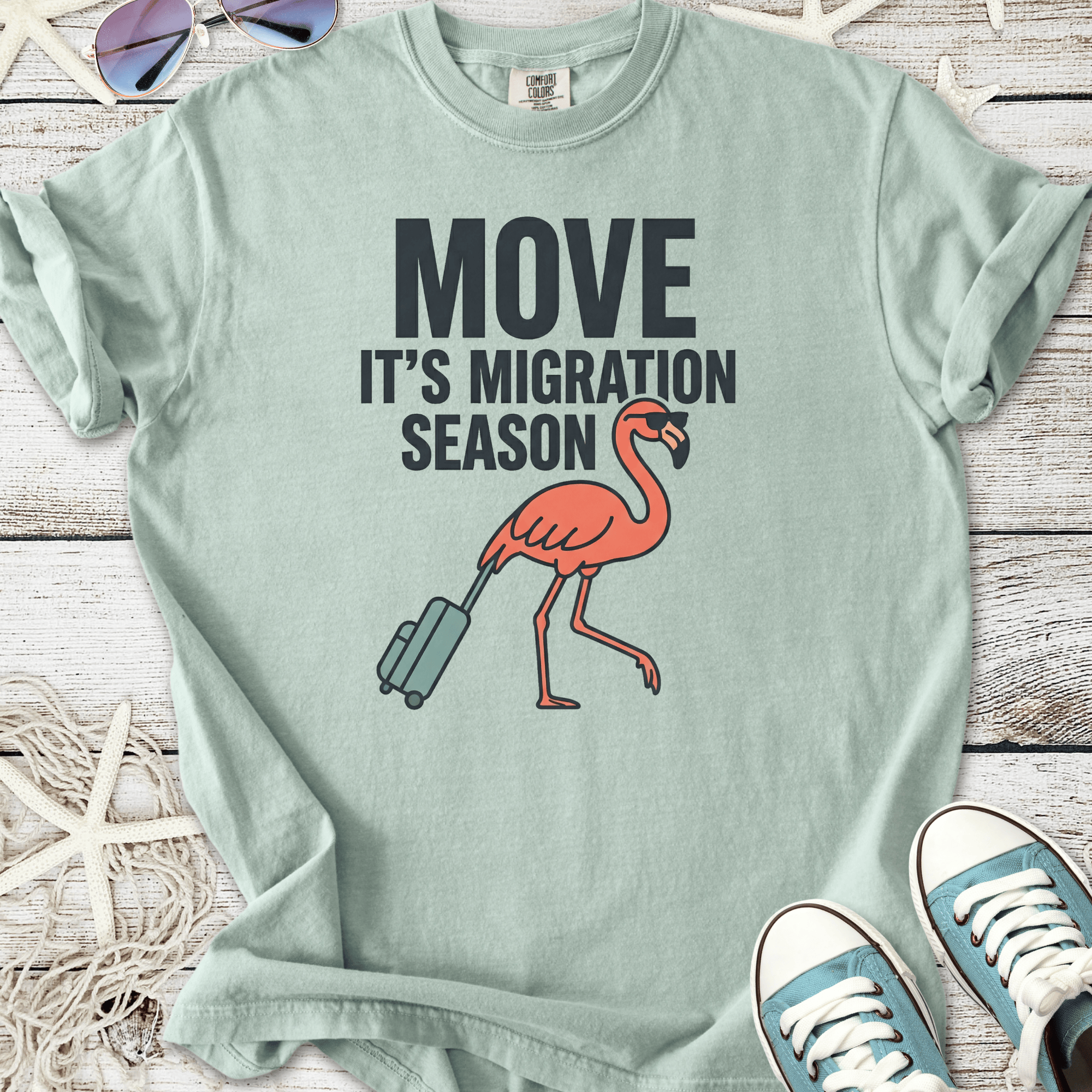 Light teal t-shirt with 'Move It's Migration Season' graphic and a pink flamingo carrying luggage.