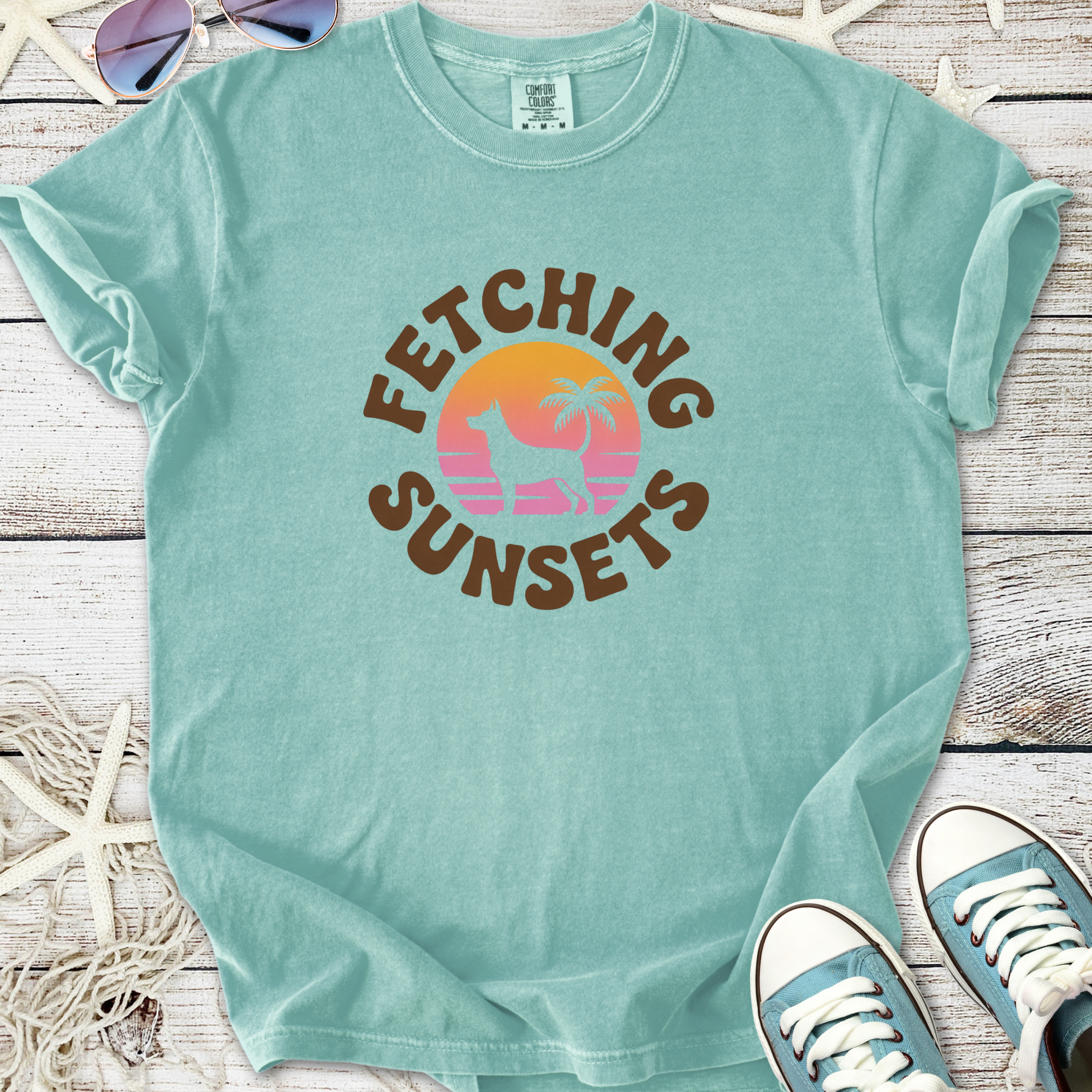 A light teal t-shirt featuring a sunset design with a dog and palm tree, perfect for casual wear.
