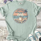 Light green t-shirt featuring 'Coffee Tastes Better with an Ocean View' graphic design.