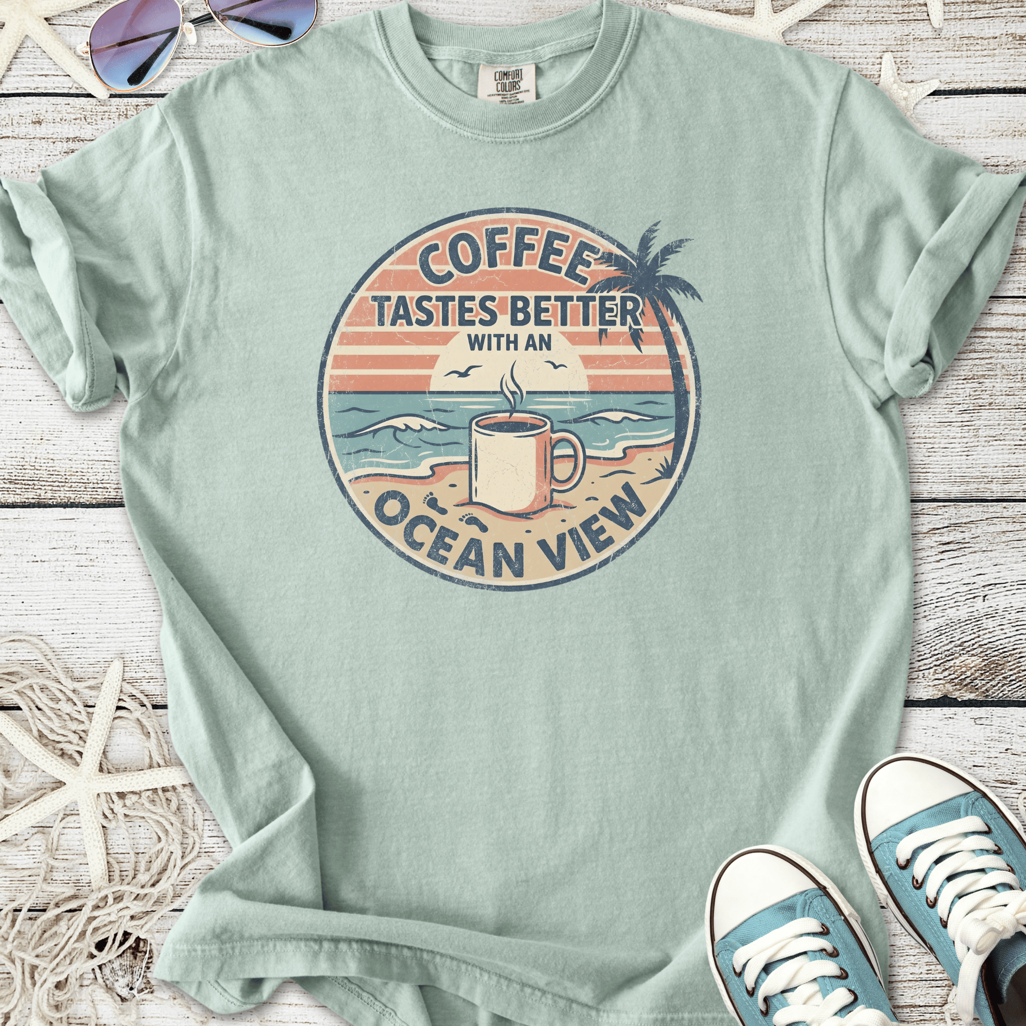 Light green t-shirt featuring 'Coffee Tastes Better with an Ocean View' graphic design.