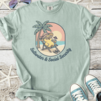 Light blue tee featuring a cute chick in sunglasses, relaxing under a palm tree with the phrase 'Saltwater & Social Security'.