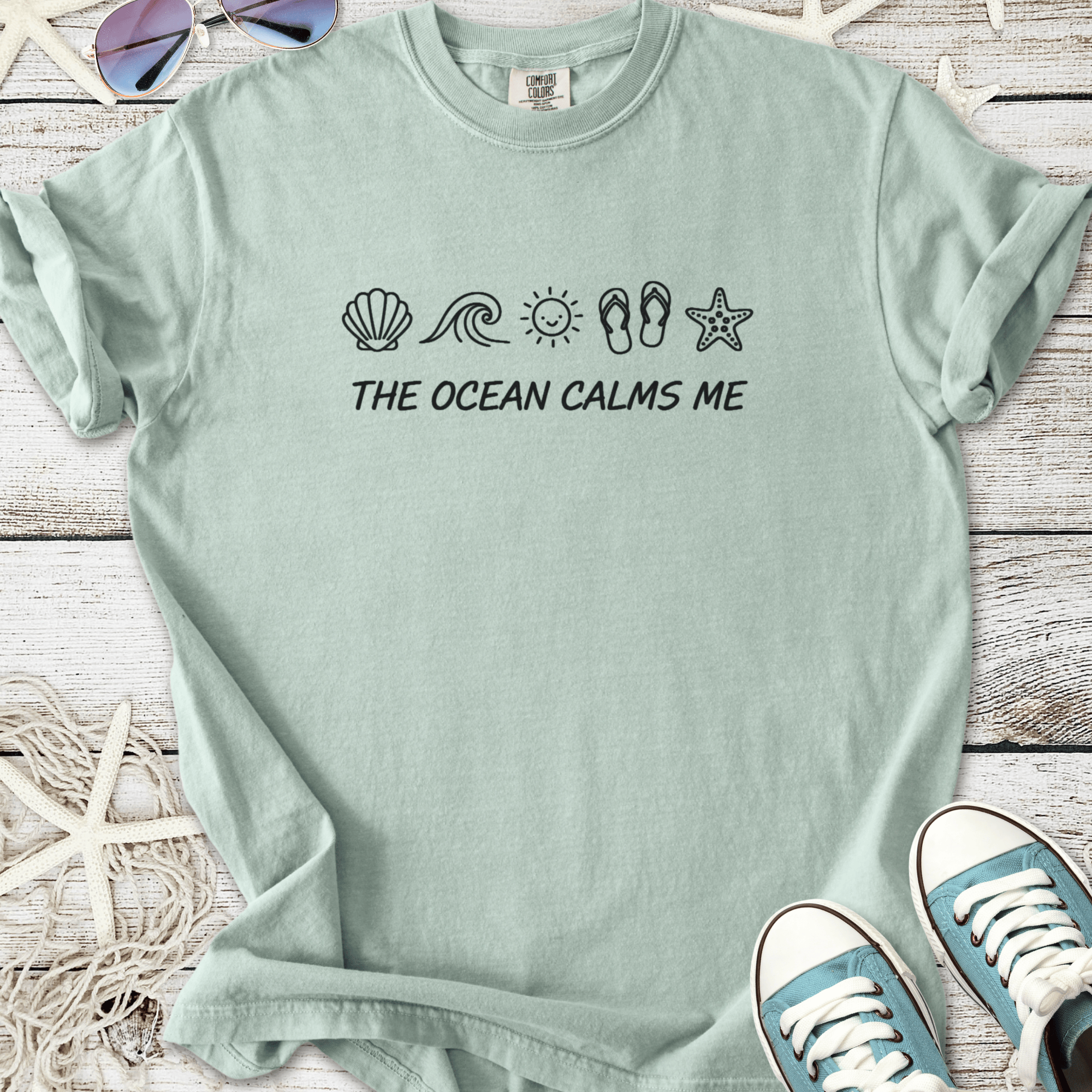 Light teal t-shirt with 'The Ocean Calms Me' print and beach-themed graphics. Comfortable cotton material.