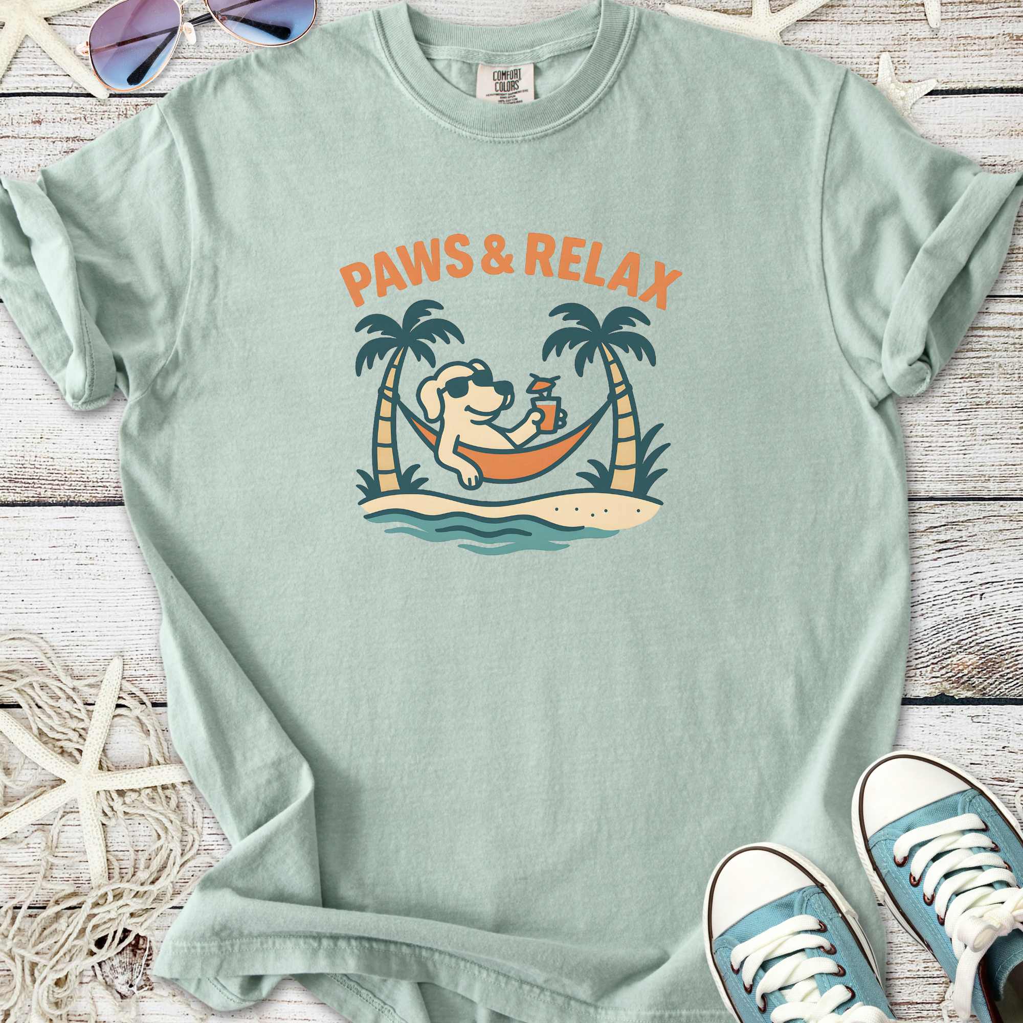 Light blue t-shirt with 'Paws & Relax' graphic, featuring a dog lounging in a hammock, tropical theme.