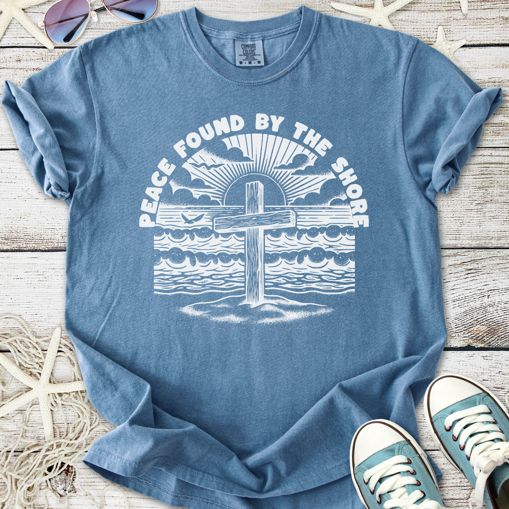Blue unisex t-shirt with 'Peace Found By The Shore' graphic, featuring a cross, waves, and sun design.