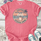 Pink graphic tee featuring 'Coffee Tastes Better with an Ocean View' design, perfect for casual wear.