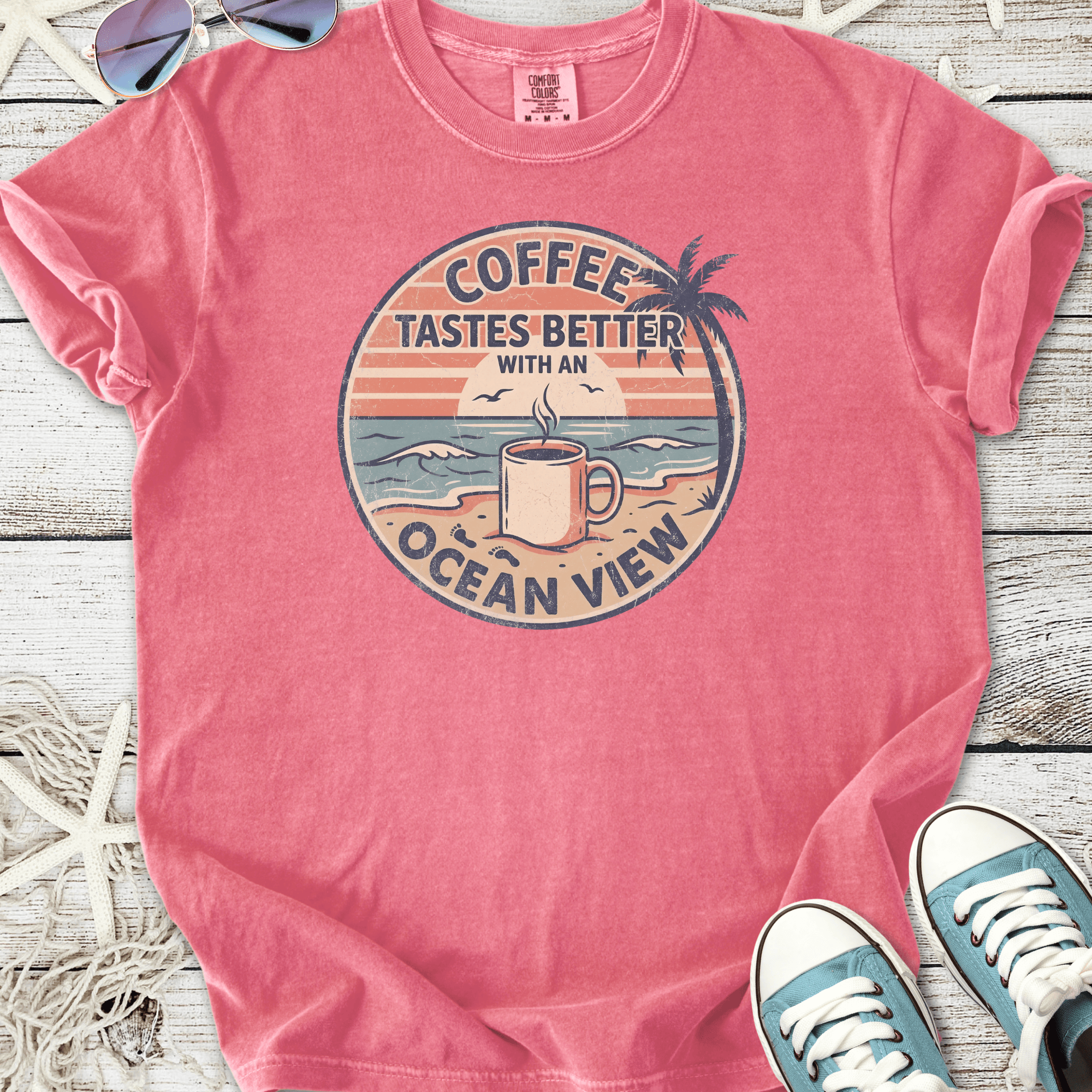 Pink graphic tee featuring 'Coffee Tastes Better with an Ocean View' design, perfect for casual wear.
