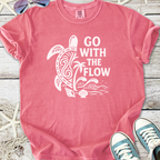 Pink graphic tee with 'Go With The Flow' design featuring turtle, palm tree, and ocean waves.