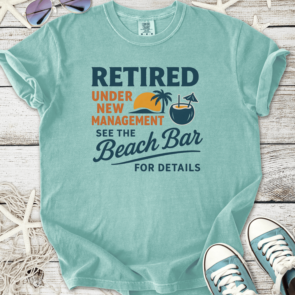 Light blue t-shirt with 'Retired Under New Management' graphic, featuring beach elements and relaxed style.