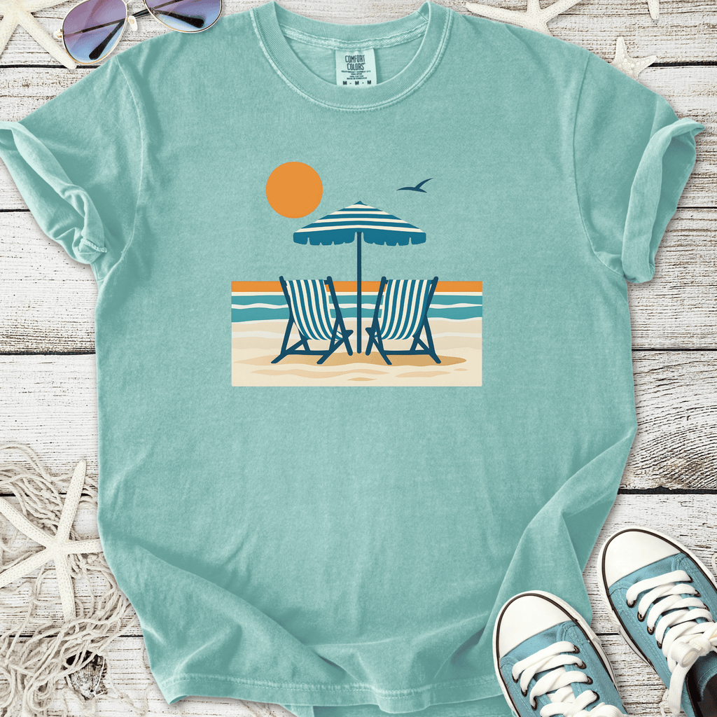 Light blue beach-themed t-shirt featuring deck chairs, umbrella, and sun graphic design.