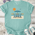 Light blue beach-themed t-shirt featuring deck chairs, umbrella, and sun graphic design.