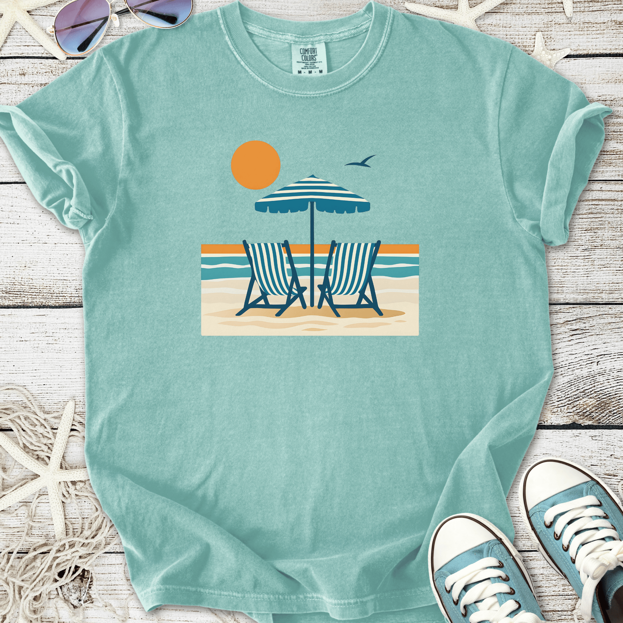Light blue beach-themed t-shirt featuring deck chairs, umbrella, and sun graphic design.