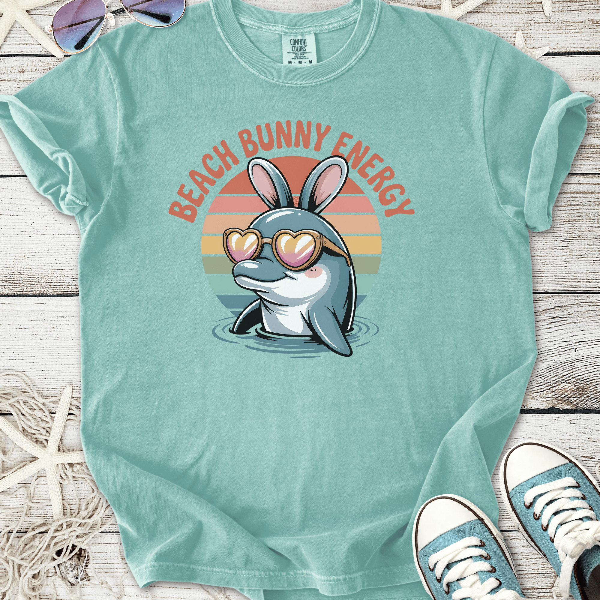 Light teal t-shirt featuring a cartoon dolphin with bunny ears and heart sunglasses, labeled 'Beach Bunny Energy'.