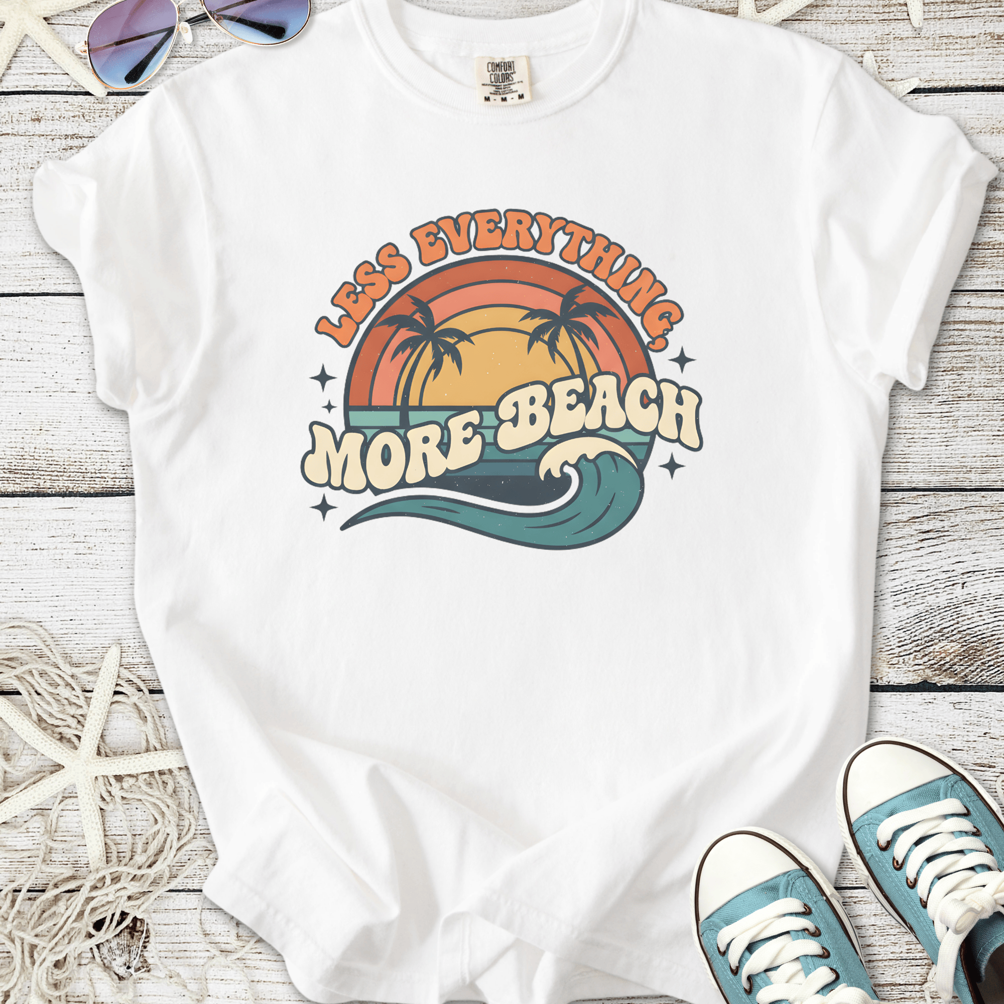 White graphic t-shirt featuring 'Less Everything, More Beach' with palm trees and waves design.