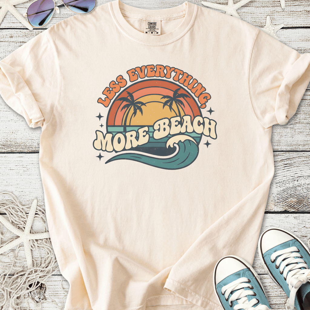 Cream-colored t-shirt with 'Less Everything, More Beach' graphic, featuring palm trees and waves.