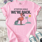 Pink graphic t-shirt featuring a flamingo with luggage, stating 'WE'RE BACK.' Fun summer casual wear.