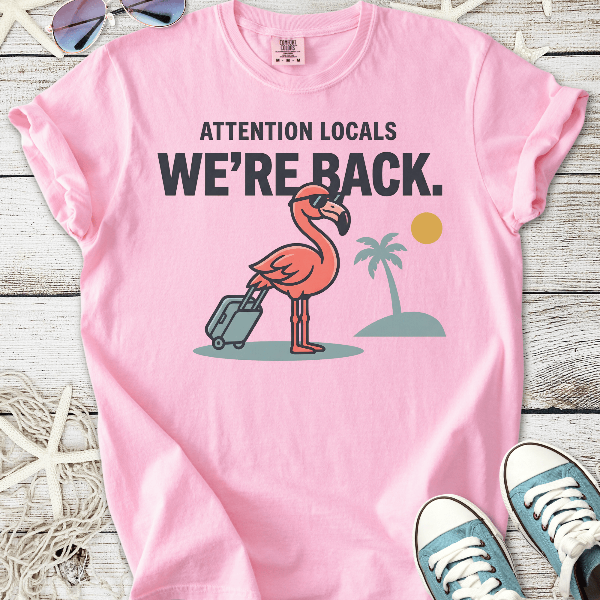 Pink graphic t-shirt featuring a flamingo with luggage, stating 'WE'RE BACK.' Fun summer casual wear.