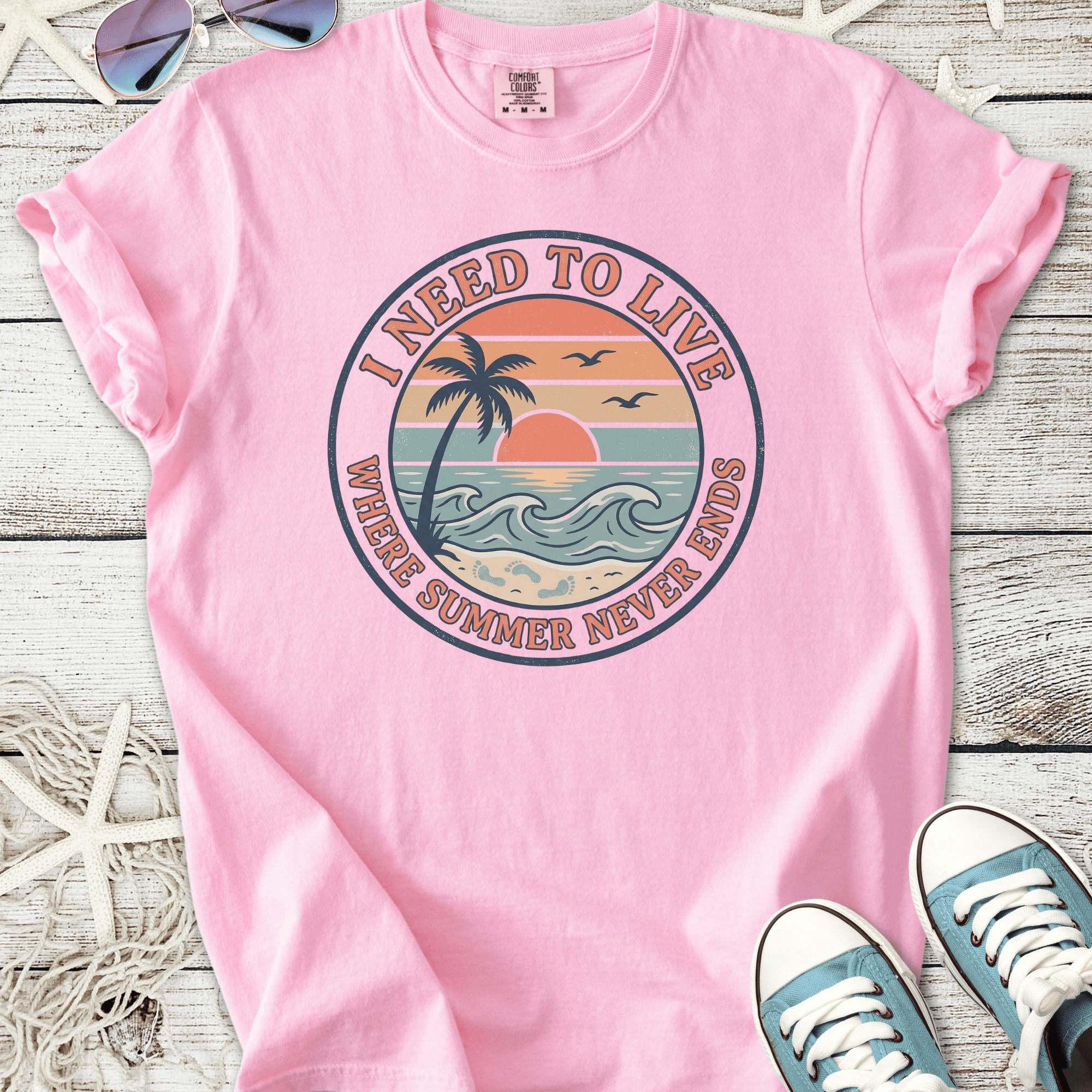 Light pink t-shirt with 'I Need To Live' graphic, featuring palm trees and sunset design, perfect for summer.