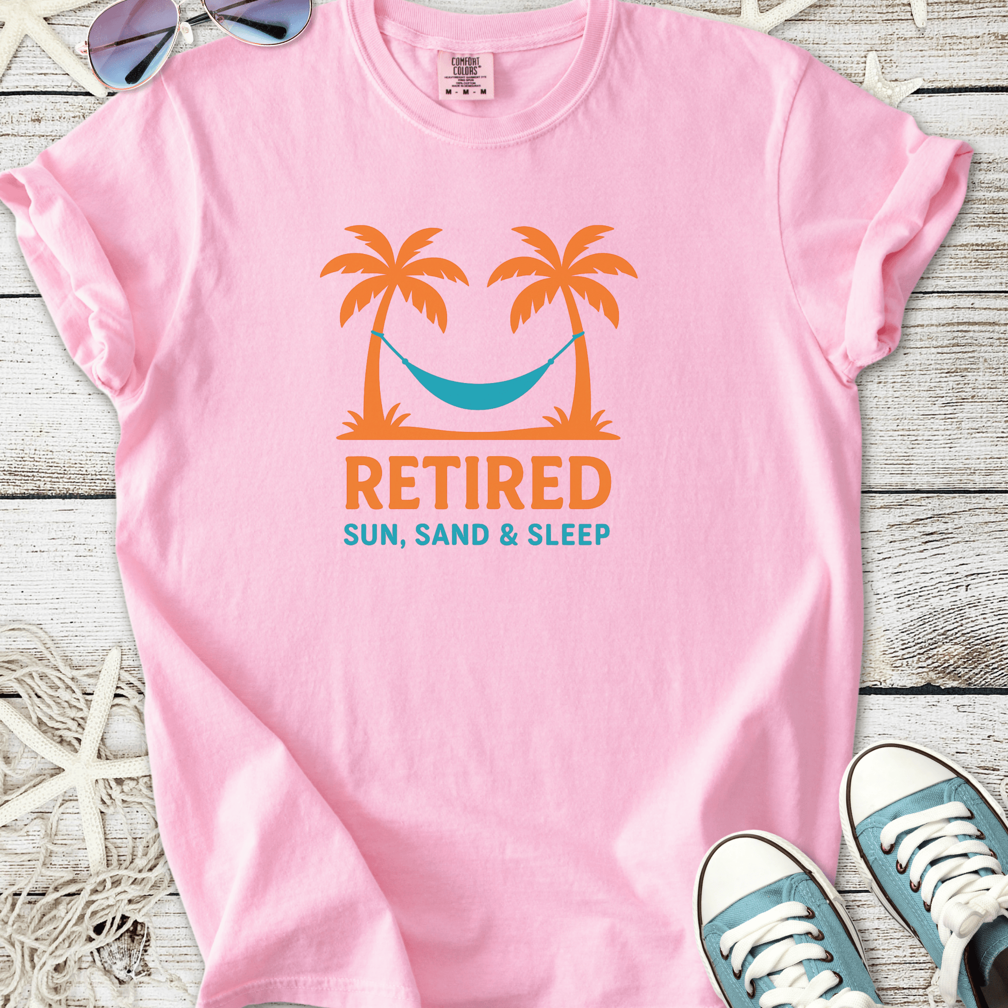 Pink cotton t-shirt with 'Retired, Sun, Sand & Sleep' graphic featuring palm trees and a hammock.