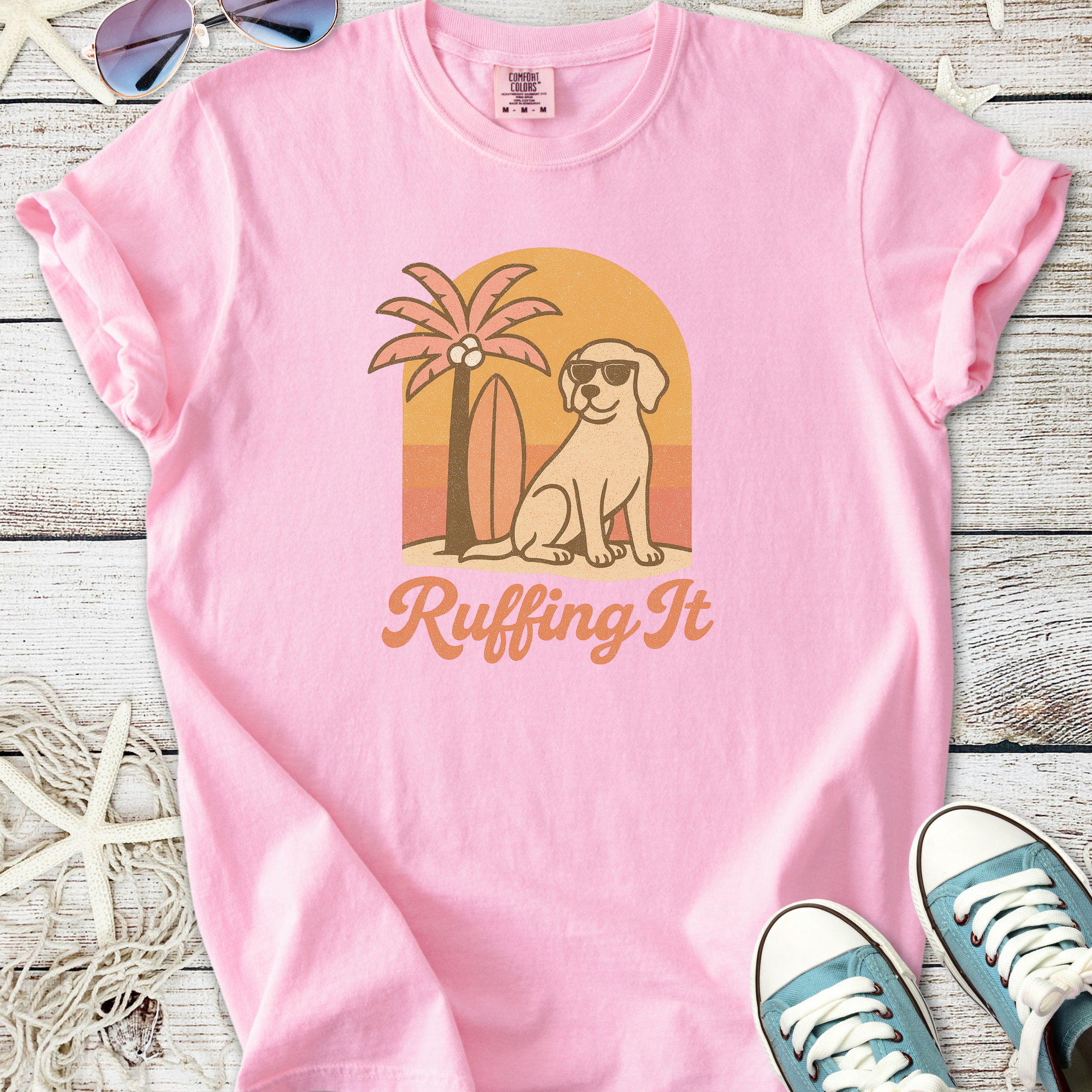 Pink t-shirt featuring a cartoon dog and palm tree design with 'Ruffing It' text, perfect for casual wear.