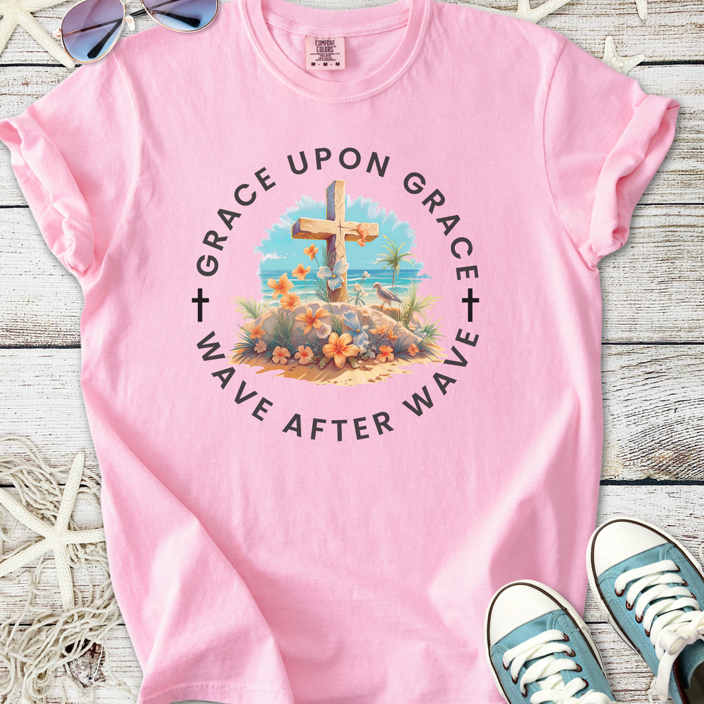 Pink graphic t-shirt featuring a beach scene with a cross and floral design, perfect for casual wear.