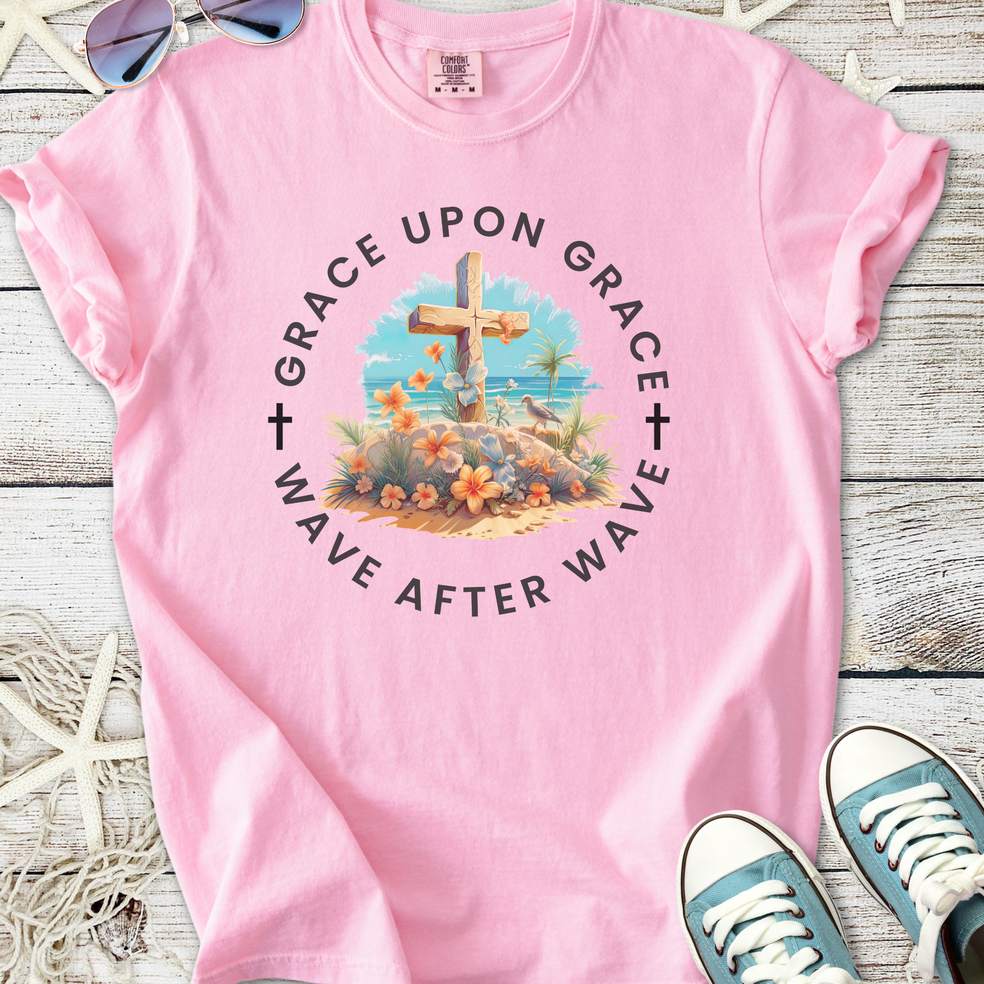 Pink graphic t-shirt featuring a beach scene with a cross and floral design, perfect for casual wear.