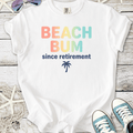 White t-shirt with 'BEACH BUM since retirement' text and palm tree design, ideal for beach lovers.