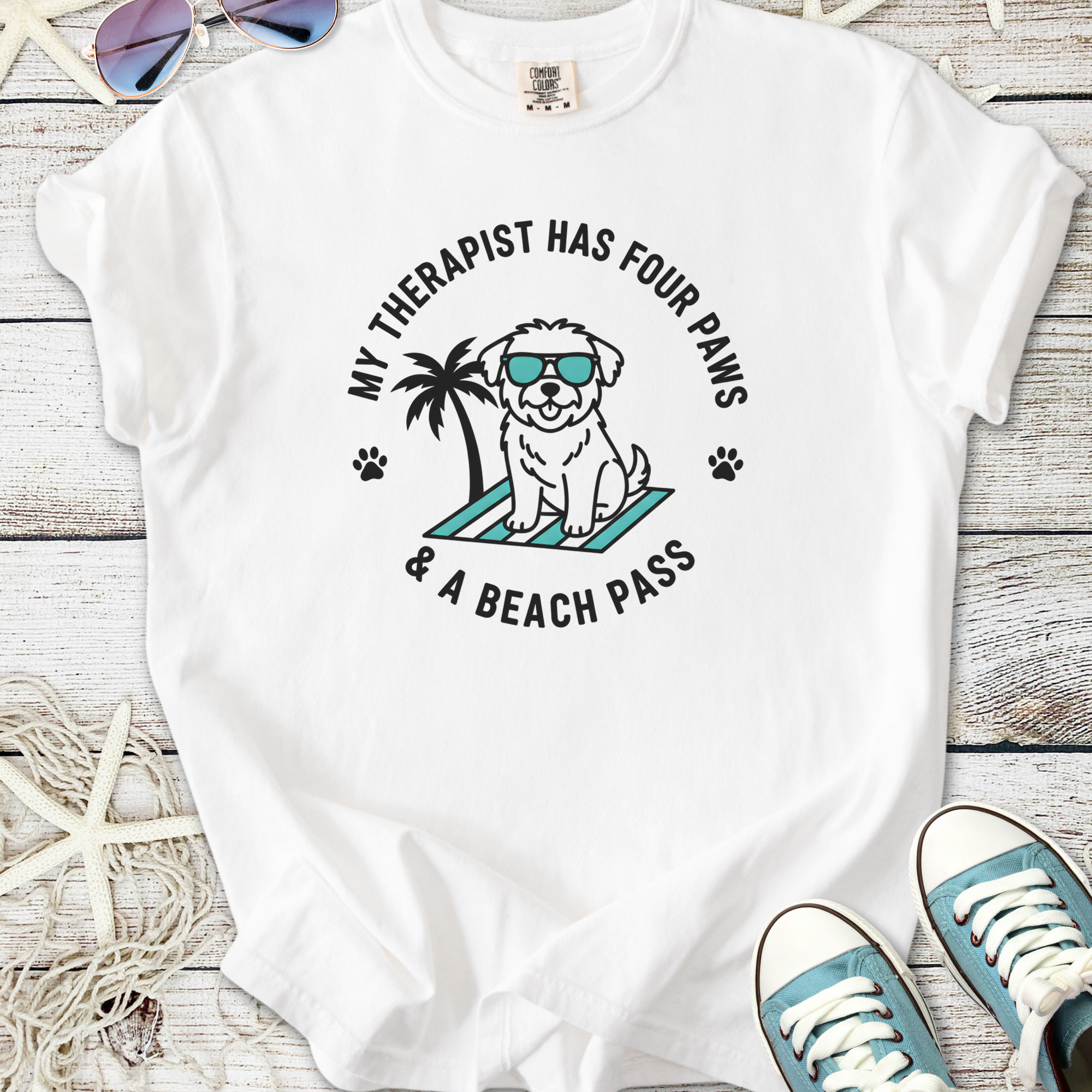 White t-shirt featuring a cute dog graphic, text "My therapist has four paws & a beach pass", perfect for dog lovers.