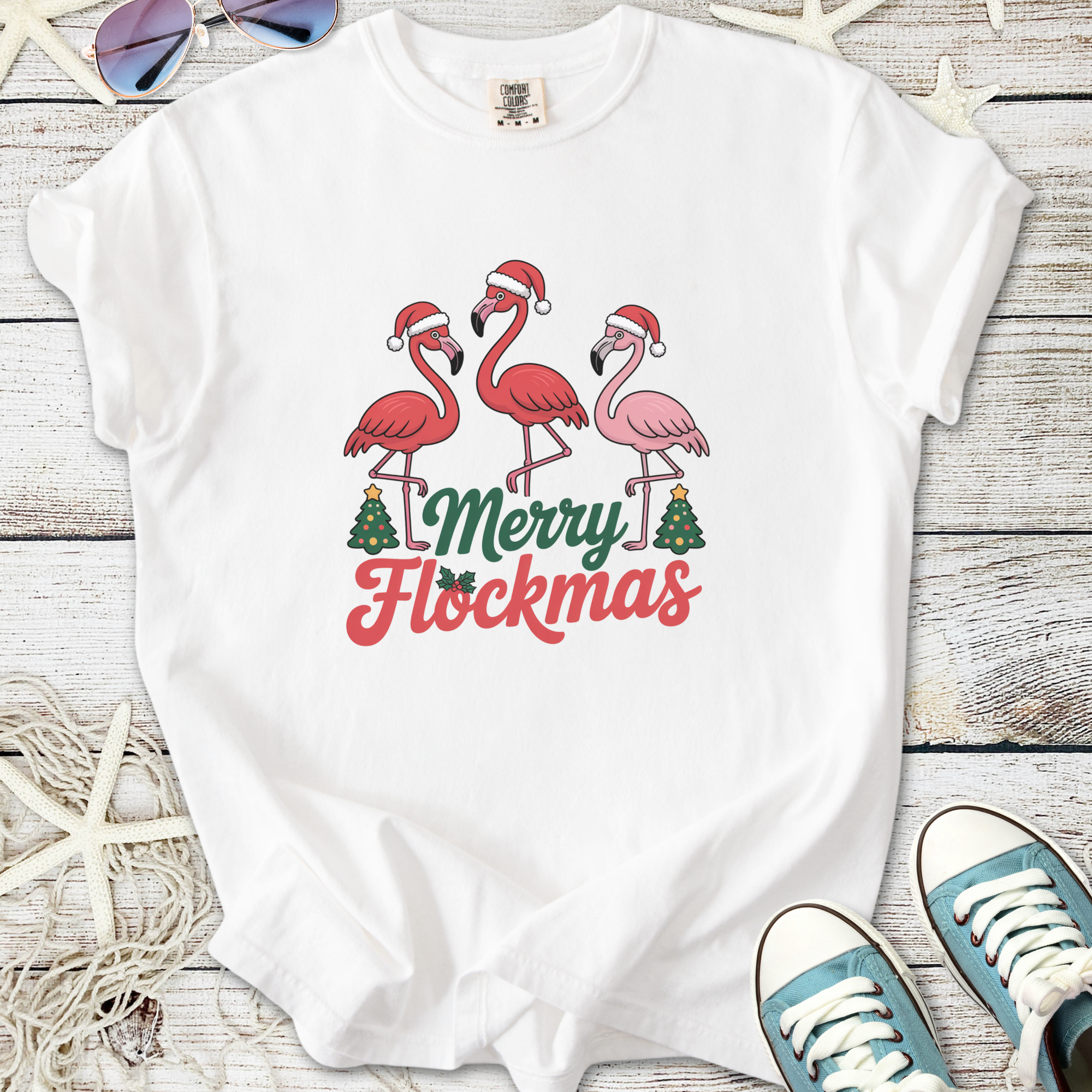 White t-shirt featuring festive flamingos with Santa hats and 'Merry Flockmas' text in vibrant colors.