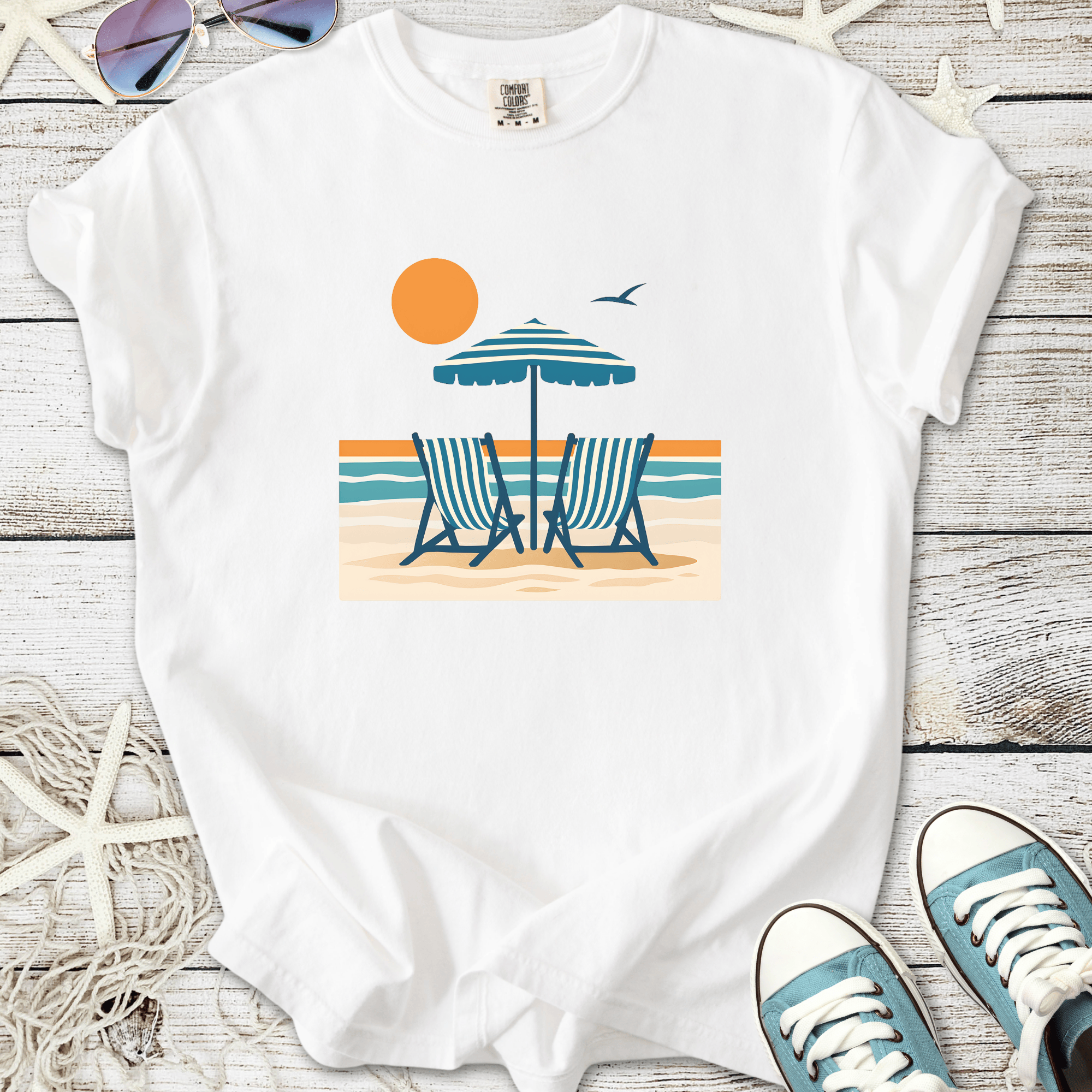 White t-shirt featuring beach scene with chairs, umbrella, sun, and ocean design. Perfect for summer vibes.