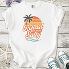 White t-shirt featuring 'Retired but Still Salty' graphic with palm tree and sunset design.