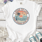 White t-shirt with 'I Need to Live' graphic featuring sunset, palm tree, and ocean waves design.