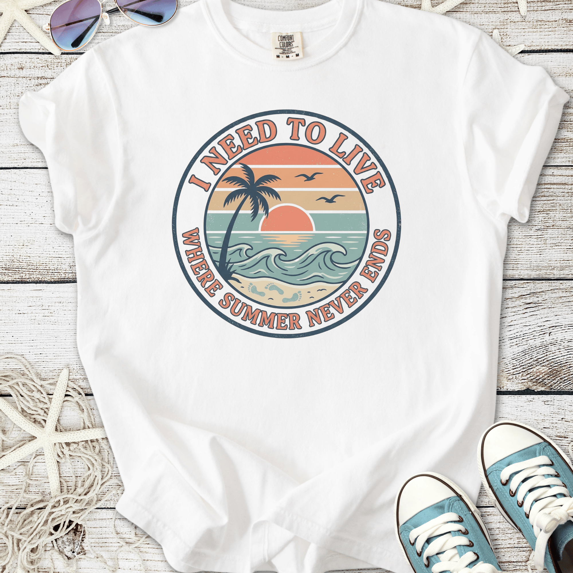 White t-shirt with 'I Need to Live' graphic featuring sunset, palm tree, and ocean waves design.