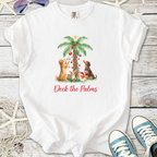 White t-shirt featuring dogs decorating a palm tree, with 'Deck the Palms' text, perfect for holiday wear.
