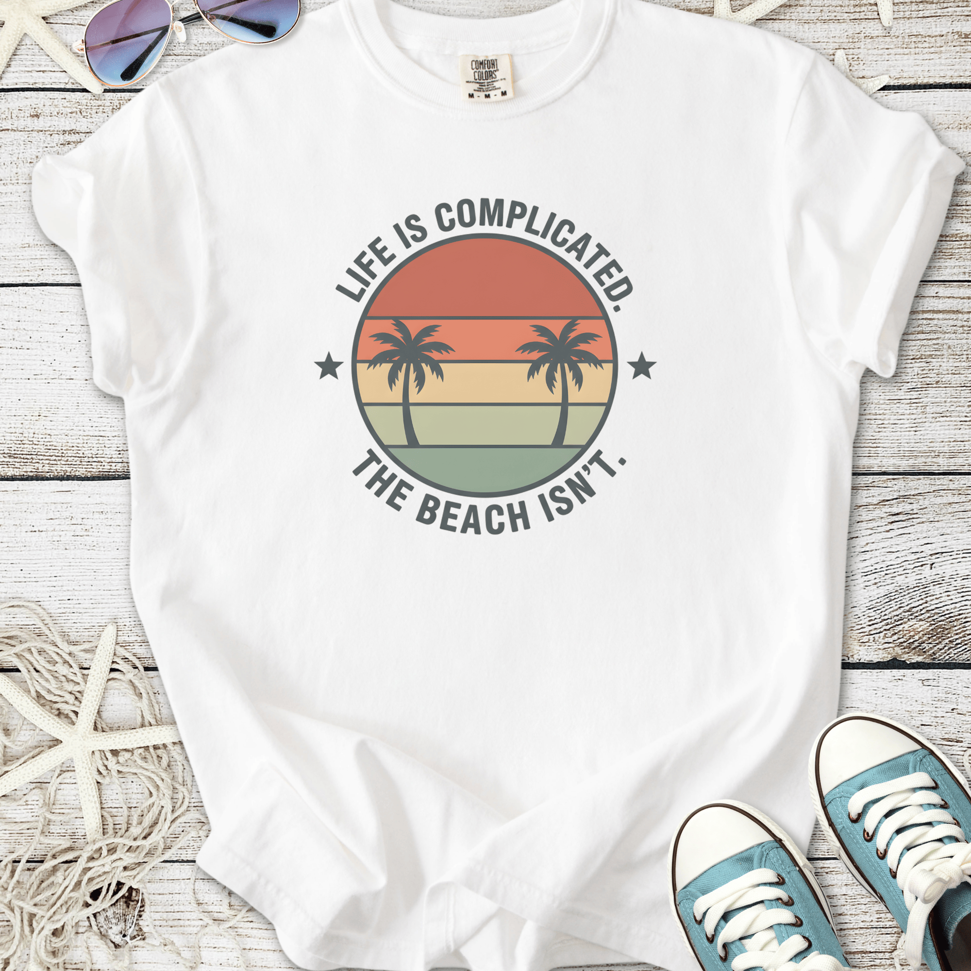 White t-shirt with palm tree graphic and 'Life is Complicated. The Beach Isn’t.' slogan.