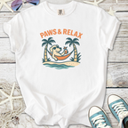 White t-shirt featuring a dog relaxing in a hammock with palm trees, text reads 'Paws & Relax'.