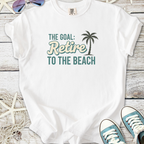 White t-shirt with palm tree graphic and text 'The Goal: Retire to the Beach' on wooden background.