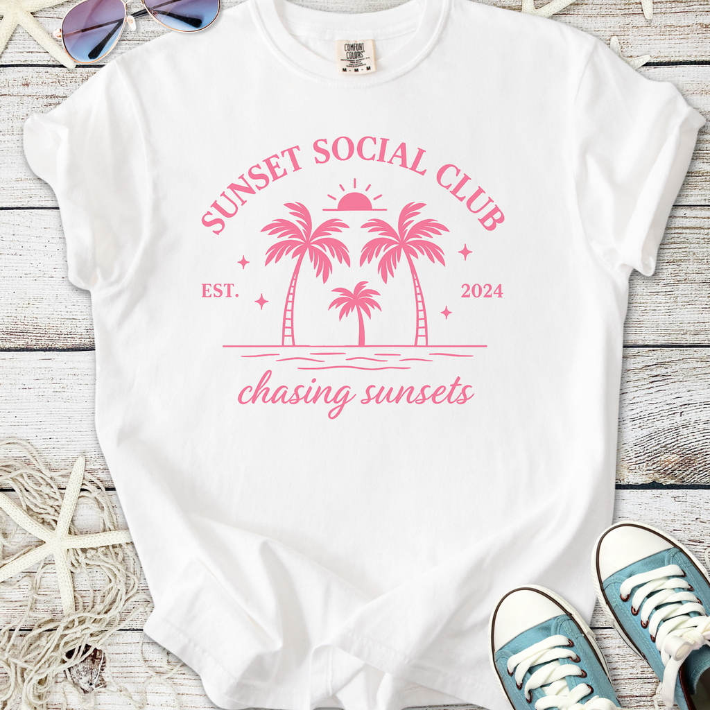 White t-shirt with pink 'Sunset Social Club' design featuring palm trees and sunset, perfect for summer outings.