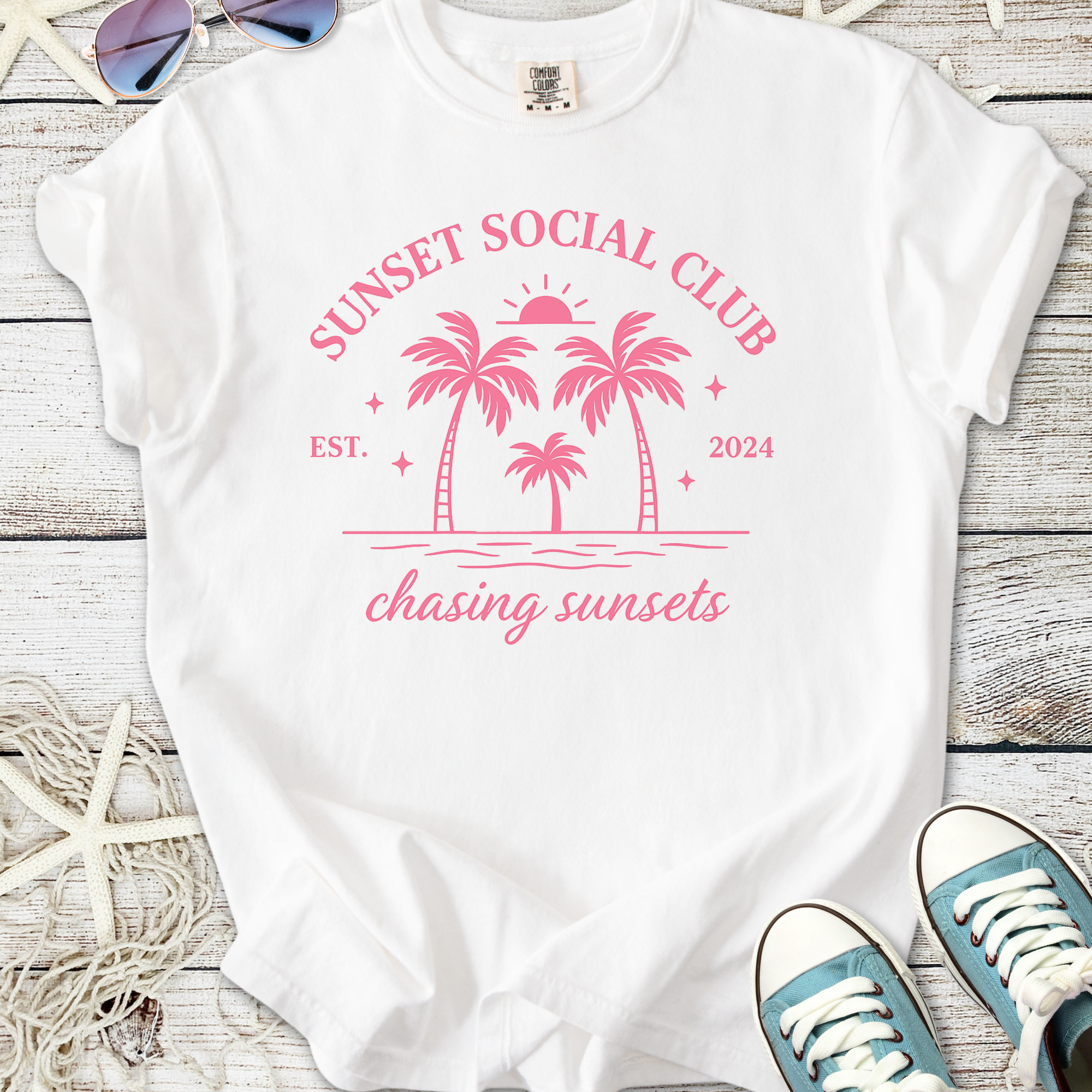White t-shirt with pink 'Sunset Social Club' design featuring palm trees and sunset, perfect for summer outings.